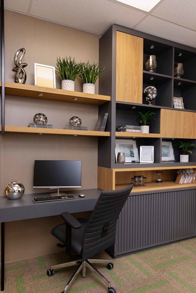 Este projeto de Home Office possui uma mesa embutida ao armário e prateleiras. A mesa do computador possui acabamento em tom preto, sobreposta por prateleiras de madeira natural. O móvel está decorado com peças de metal, vasos de plantas, porta-retratos e livros.